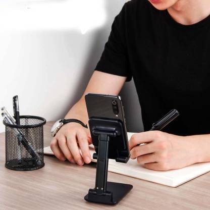 A person is using a black mobile stand with adjustable design on a desk to hold a smartphone. The stand is supporting a phone while the person is writing on a notebook.